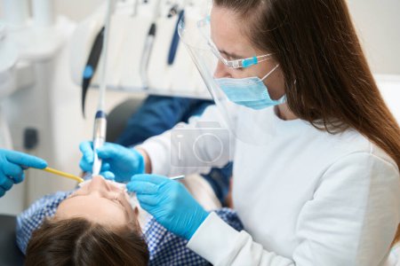 Foto de Mujer dentista realiza procedimiento higiénico para el paciente joven, un asistente ayuda al médico en su trabajo, utilizan un endomotor - Imagen libre de derechos