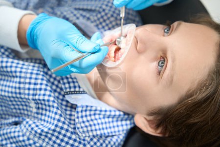 Téléchargez les photos : Dentiste vérifie l'état des dents de garçon dans des gants de protection, le médecin utilise des outils spéciaux - en image libre de droit