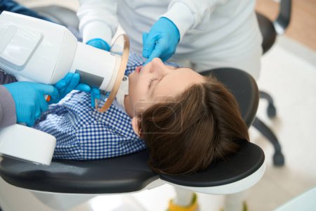 Téléchargez les photos : Adolescent a une radiographie dentaire dans le cabinet dentaire, la procédure est effectuée par des spécialistes en uniformes médicaux - en image libre de droit