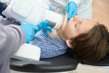 Foto de El niño recibe una radiografía del diente en el consultorio dental, el procedimiento es llevado a cabo por especialistas en uniformes médicos y guantes - Imagen libre de derechos