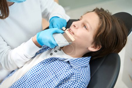 Foto de Especialista en una clínica dental coincide con los colores de un diente a un adolescente con una camisa a cuadros - Imagen libre de derechos