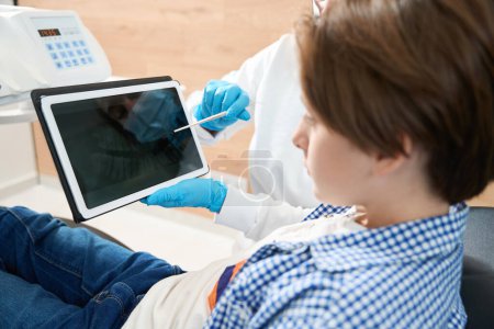 Téléchargez les photos : Dentiste consultant un jeune patient au cabinet dentaire, le médecin utilise une tablette et un localisateur de sommet - en image libre de droit