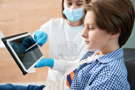 Téléchargez les photos : Femme dentiste en uniforme et gants montre une image dentaire sur un presse-papiers à une jeune patiente - en image libre de droit