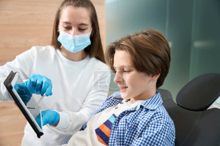 Garçon à un rendez-vous chez le dentiste, une femme dentiste lui montre une photo sur une tablette et lui conseille