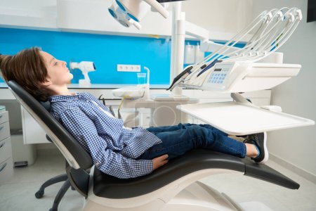 Konzentrierter Junge im karierten Hemd sitzt ruhig im Zahnarztstuhl, Spezialgeräte liegen herum