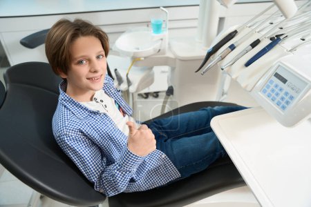 Téléchargez les photos : Enfant assis dans la chaise dentaire, il sourit et montre ok - en image libre de droit