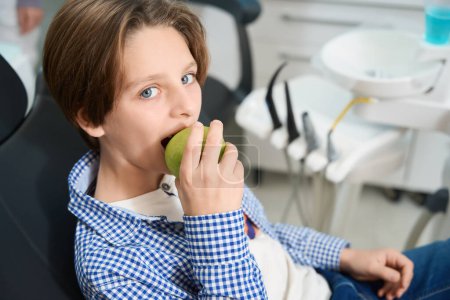 Téléchargez les photos : Garçon assis dans la chaise du dentiste sur le fond d'outils spéciaux, il mange une pomme - en image libre de droit