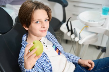 Téléchargez les photos : Un garçon souriant assis dans une chaise de dentiste sur fond d'outils spéciaux, il a une pomme dans les mains - en image libre de droit