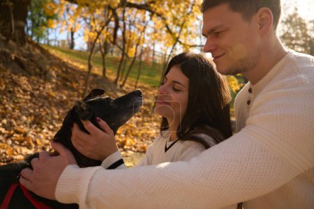 Foto de Guy y su novia están sonriendo y acariciando al perro, están paseando en el parque de otoño - Imagen libre de derechos