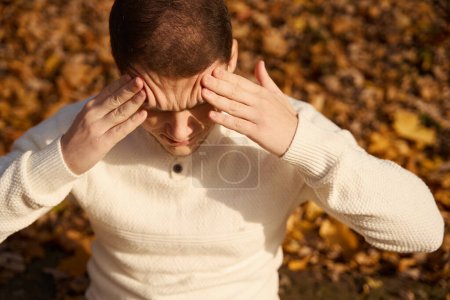 Téléchargez les photos : L'homme souffre et tient son front, il est assis dans un parc d'automne - en image libre de droit