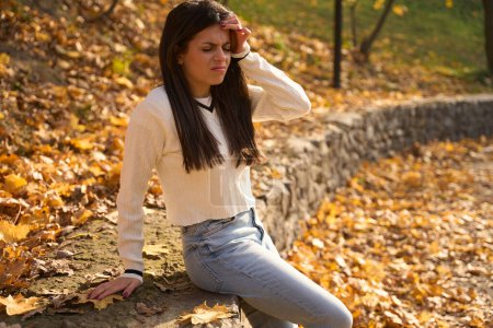 Téléchargez les photos : Femme froncée des sourcils s'assoit sur un parapet et tient sa tête, feuilles d'automne se trouvent autour - en image libre de droit