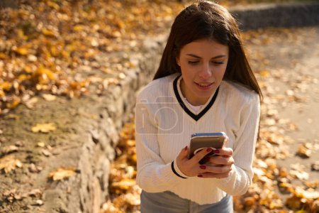 Téléchargez les photos : Intéressé jeune femme tenant un téléphone dans ses mains, feuilles d'automne autour - en image libre de droit