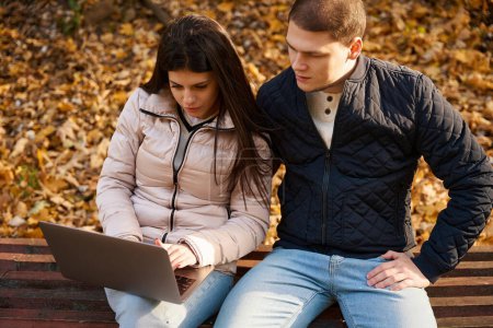 Foto de Jóvenes enfocados están leyendo algo en un portátil, un tipo con una novia están sentados en el parque - Imagen libre de derechos