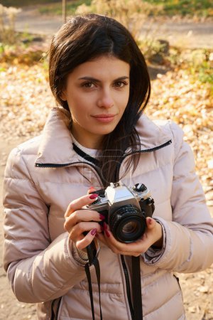 Photo for Beautiful dark-haired woman holds a camera in her hands, she stands in a city park - Royalty Free Image