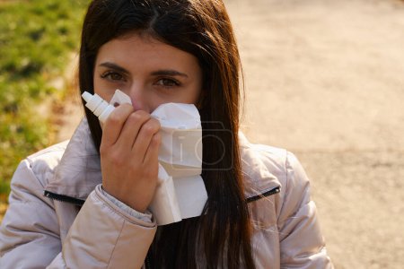 Photo for Sick woman uses a paper handkerchief and a spray for a cold, she is in the park - Royalty Free Image