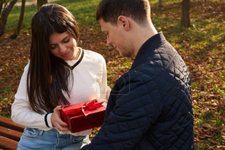 Photo for Pretty young woman accepts a gift box from her boyfriend, they are relaxing on a bench in the park - Royalty Free Image