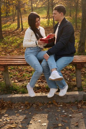 Photo for Young woman holds gift box with bow in her hands, she and her boyfriend are sitting on bench in park - Royalty Free Image