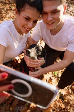 Foto de Feliz estudiante pareja tomar selfie con su perro en el contexto de la naturaleza de otoño, sonríen a la cámara del teléfono - Imagen libre de derechos