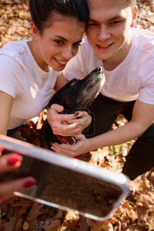 Téléchargez les photos : Jeune couple mignon prend selfie avec leur chien sur le fond de feuilles tombées, ils sourient à la caméra du téléphone - en image libre de droit