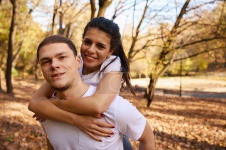 Athletic guy rides his joyful girlfriend on backs, they frolic in a beautiful park