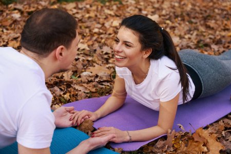 Photo for Happy boyfriend and his girlfriend are holding hands and looking at each other, they are lying on karimats in autumn park - Royalty Free Image