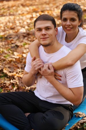 Photo for Guy with slight unshaven and his girlfriend, hugging each other, are sitting on carimat in the park, fallen leaves around - Royalty Free Image
