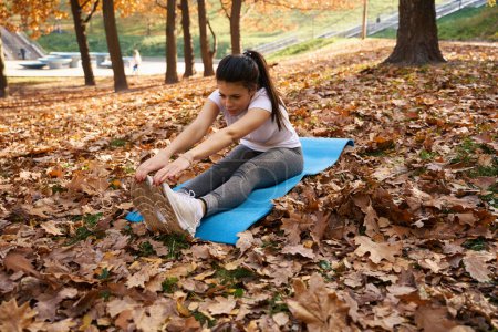 Photo for Woman stretching on a blue carimate in a city park among autumn foliage - Royalty Free Image