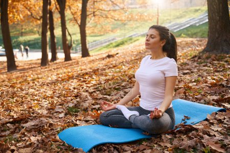 Photo for Calm woman with closed eyes sits in a park in a lotus position among yellow fallen leaves - Royalty Free Image