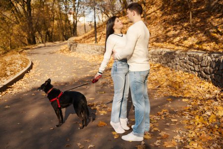 Foto de Un tipo con un suéter ligero abraza a su novia en un callejón de paisaje, una mujer mantiene a un perro con una correa - Imagen libre de derechos