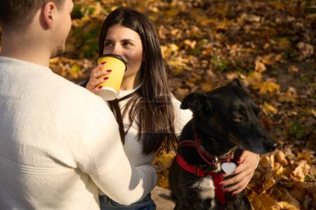 Foto de Hermosa mujer joven está tomando café y charlando con su novio en el parque de otoño, que están paseando a su amado perro - Imagen libre de derechos