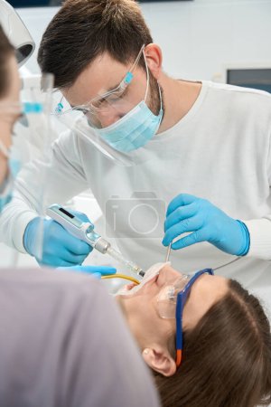 Foto de Médico en guantes de nitrilo desechables inyectando anestésico al cliente con pistola de jeringa dental - Imagen libre de derechos