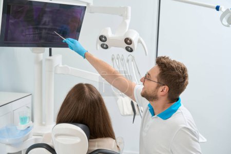 Téléchargez les photos : Dentiste pointant vers la radiographie dentaire virtuelle à l'écran de l'ordinateur à la patiente - en image libre de droit