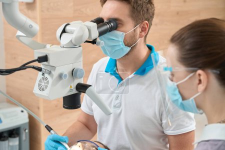 Photo for Qualified stomatologist carrying out dental procedure on patient under microscope assisted by female nurse - Royalty Free Image