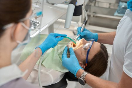 Foto de Estomatólogo extirpando sarro entre dientes frontales de mujer joven asistida por enfermera - Imagen libre de derechos
