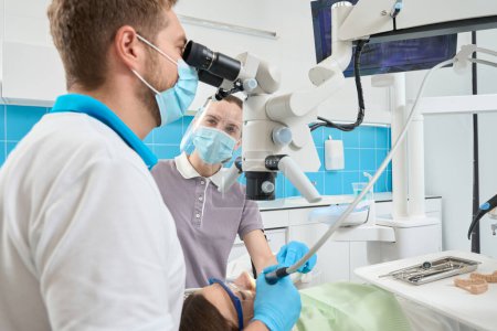 Foto de Asistente experimentado ayudando al estomatólogo durante el procedimiento médico realizado en el cliente bajo el microscopio - Imagen libre de derechos