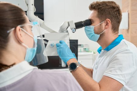 Foto de Estomatólogo mirando a través del ocular del microscopio dental y girando la perilla de enfoque supervisado en presencia de la enfermera - Imagen libre de derechos
