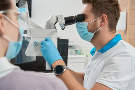 Foto de Dentista mirando a través del ocular del microscopio y girando la perilla de enfoque supervisado por la enfermera - Imagen libre de derechos