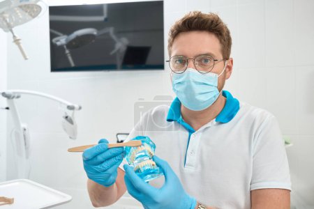 Foto de Estomatólogo en mascarilla desechable y guantes de nitrilo cepillado modelo dientes con cepillo de dientes - Imagen libre de derechos