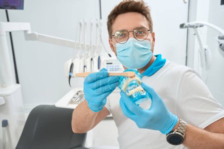 Photo for Dental hygienist in face mask and nitrile gloves brushing upper front teeth on transparent human jaw model with toothbrush - Royalty Free Image