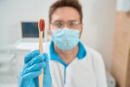 Foto de Retrato borroso del higienista dental en mascarilla facial y guante de nitrilo sosteniendo el cepillo de dientes ante la cámara - Imagen libre de derechos