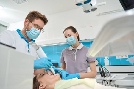 Téléchargez les photos : Dentiste focalisé scannant les dents supérieures de la patiente avec scanner intra-buccal supervisé par un assistant - en image libre de droit