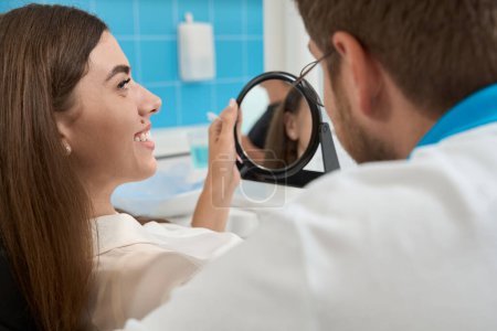 Téléchargez les photos : Agréable patiente assise dans une chaise tenant un miroir de main et regardant son dentiste - en image libre de droit