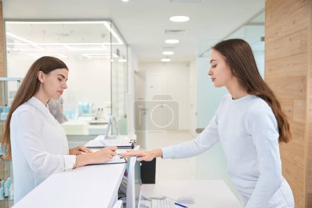 Foto de Cliente femenina joven enfocada seria llenando formulario de consentimiento en presencia de recepcionista - Imagen libre de derechos