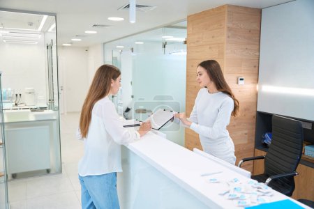 Female patient filling in consent form in hands of young clinic receptionist