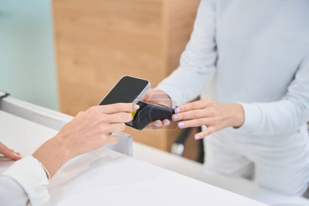 Téléchargez les photos : Photo recadrée d'une cliente tenant un téléphone portable sur un terminal PDV entre les mains d'une réceptionniste - en image libre de droit