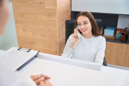 Smiling friendly secretary seated at reception desk looking at client during phone conversation