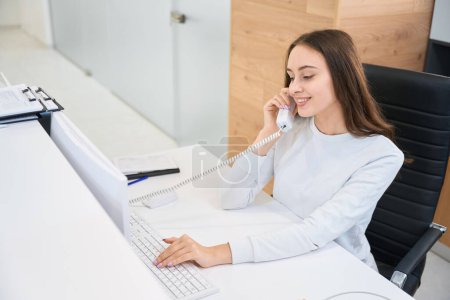 Téléchargez les photos : Sourire heureux jeune réceptionniste femelle tapant sur ordinateur de bureau pendant la conversation téléphonique - en image libre de droit