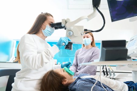 Root canal treatment for a boy in a modern dental clinic is carried out with anesthesia, under a microscope, the dentist works with an assistant
