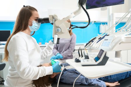 Téléchargez les photos : Femme médecin traite un patient sur un équipement moderne, utilise un microscope, et un assistant est à proximité - en image libre de droit