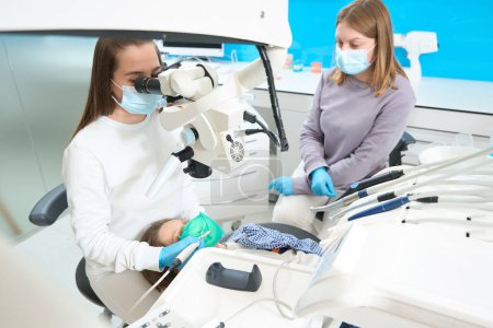 Foto de Dentista está tratando un diente de niño bajo un microscopio, ella utiliza equipos modernos, un asistente está cerca - Imagen libre de derechos